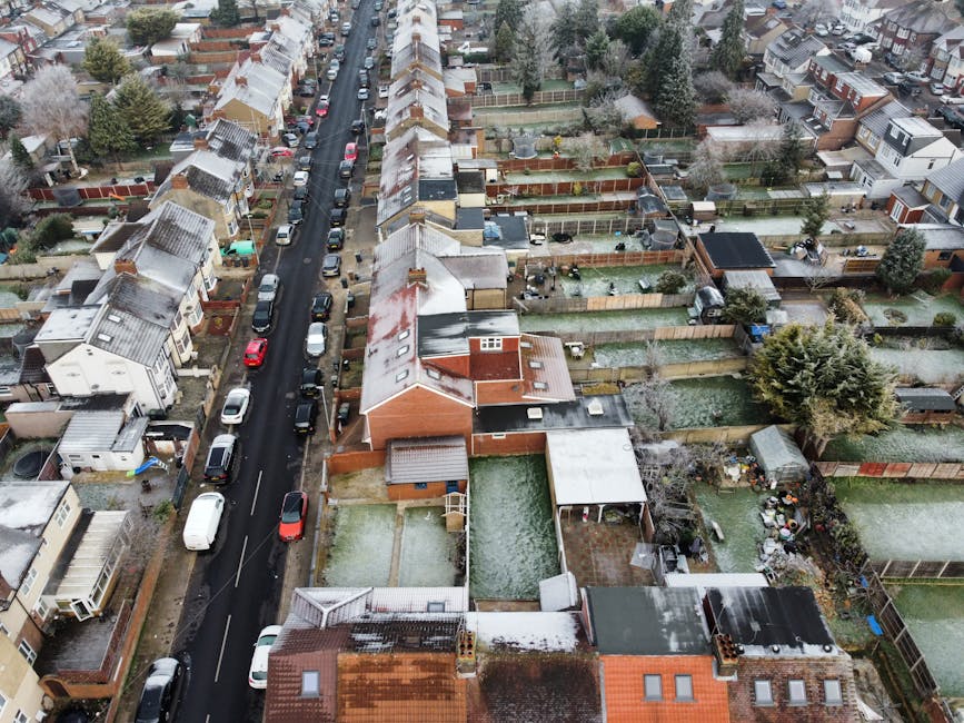 An aerial view of a residential street in West Hendon during winter, showing tightly packed terraced houses with pitched roofs, some covered in a light layer of snow. The street is lined with parked cars, including hatchbacks and vans, along both curb sides. Behind the houses, there are numerous fenced backyards, many containing garden furniture, sheds, and small trees, with some yards featuring outdoor storage units or garden decorations. The scene captures a typical urban neighbourhood with a mix of brick and timber fences, paved pathways, and garden areas. The lighting is diffuse due to overcast sky conditions, and there are no visible moving vehicles or people, suggesting a quiet moment in the day. This setting reflects a typical home environment where house removals and furniture transport may take place, highlighting the importance of careful logistics on narrow, urban streets. Man with Van West Hendon offers services that facilitate such home relocations with attention to detail in confined residential areas.