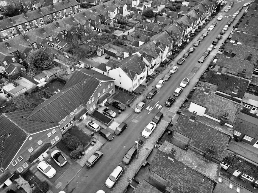 An aerial view of a residential street in West Hendon during winter, showing tightly packed terraced houses with pitched roofs, some covered in a light layer of snow. The street is lined with parked cars, including hatchbacks and vans, along both curb sides. Behind the houses, there are numerous fenced backyards, many containing garden furniture, sheds, and small trees, with some yards featuring outdoor storage units or garden decorations. The scene captures a typical urban neighbourhood with a mix of brick and timber fences, paved pathways, and garden areas. The lighting is diffuse due to overcast sky conditions, and there are no visible moving vehicles or people, suggesting a quiet moment in the day. This setting reflects a typical home environment where house removals and furniture transport may take place, highlighting the importance of careful logistics on narrow, urban streets. Man with Van West Hendon offers services that facilitate such home relocations with attention to detail in confined residential areas.