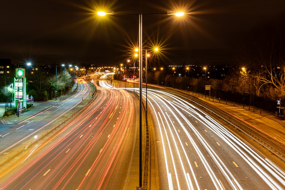 A night-time photograph of a busy multi-lane road with streaks of car headlights and taillights indicating fast-moving traffic, taken from an elevated perspective. The road is divided by a central barrier, with streetlights illuminating the scene and creating starburst effects. On the left side of the image, a petrol station sign is visible, and nearby trees and buildings are faintly illuminated in the distance. The right side features a sidewalk lined with metal railings and bare trees. This scene captures an urban setting during a typical evening, with moving vehicles providing dynamic visual cues relevant to transportation and logistics, supporting the context of home relocation and furniture transport services carried out by companies like Man with Van West Hendon.