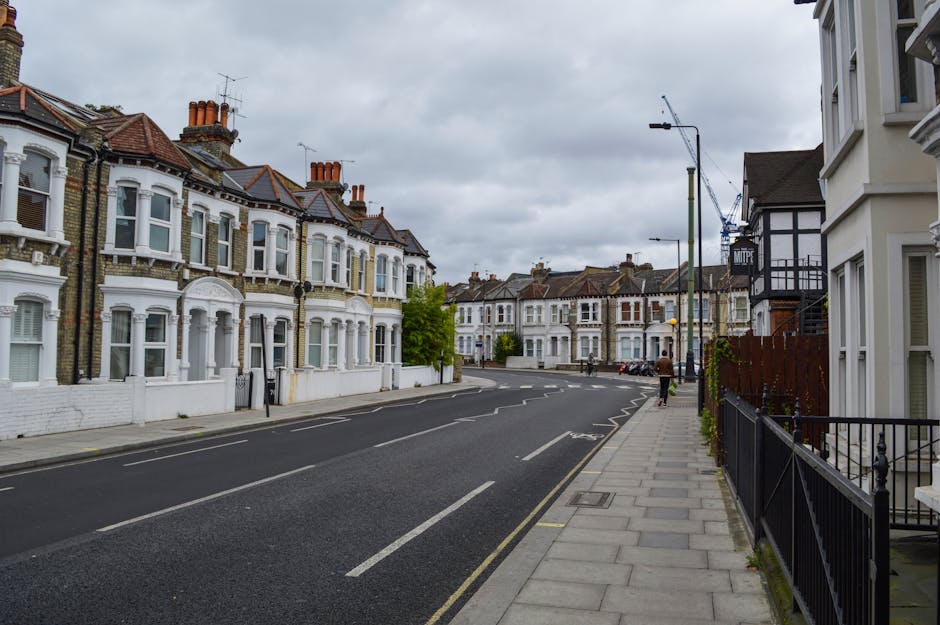 The image depicts a quiet residential street in West Hendon NW9 with rows of Victorian-style terraced houses featuring bay windows and decorative brickwork, situated on the left side of the street. The houses are primarily painted white with some brick detailing, with chimneys visible atop the roofs. The street is paved with asphalt, with clearly marked parking bays and a designated lane for moving vehicles. The sidewalk on the right side is tiled with concrete slabs, bordered by black metal railings and potted plants. In the distance, a construction crane is visible, indicating ongoing development. Overhead, the sky is cloudy, casting diffuse lighting over the scene. This setting could serve as a reference for home relocation or furniture transport logistics, aligning with services offered by Man with Van West Hendon, especially during packing and loading processes, with movement preparations potentially taking place on this street.
