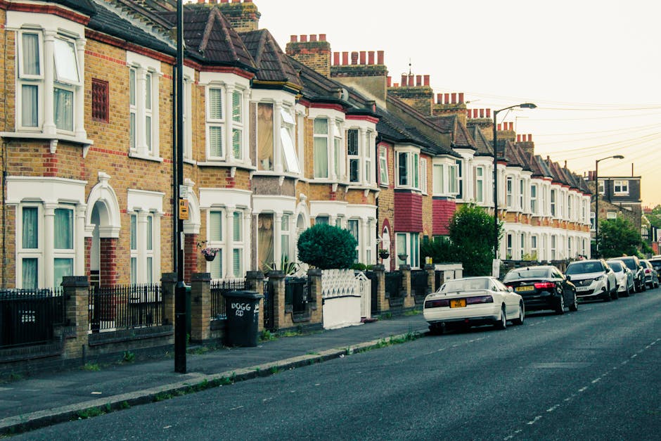 A row of Victorian-style terraced houses on West Hendon Road, featuring brick facades, white bay windows, and decorative window frames. In front of the houses, there is a pavement with a black metal fence and small front gardens containing shrubs and plants. Several vehicles, including a white car and black cars, are parked along the street. A moving truck or van is not visible, but the setting suggests a residential area where home relocation or furniture transport might occur. The scene is captured during daylight hours with overcast skies, illustrating typical urban house frontage suitable for house removals and moving services by Man with Van West Hendon.