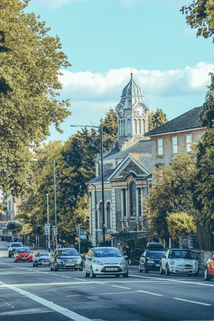 A street scene in West Hendon featuring a row of parked cars along the curb, with various models and colours, in front of a historic building with a clock tower. The building has architectural details including arched windows, decorative brickwork, and a peaked roof, partially obscured by surrounding leafy trees. Nearby streetlights and pavement are visible, highlighting an urban environment suitable for home relocation or furniture transport. The sky above is clear with a few clouds, and natural light illuminates the scene, indicating daytime. This setting exemplifies typical elements involved in house removals and moving logistics, such as vehicle parking, urban street layout, and accessible surroundings, with [COMPANY_NAME] often assisting with these types of services in the West Hendon area.