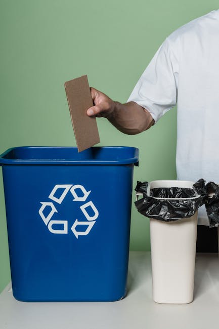 A person wearing a white shirt holds a cardboard piece over a large blue recycling bin with a white recycling symbol printed on it, positioned on a flat surface. Next to the blue bin is a smaller white container lined with a black plastic bag, partially filled with waste materials. The background features a plain light green wall, and the scene appears to be set indoors, likely as part of house relocation or packing and moving activities. The image depicts waste separation and disposal procedures related to house removals, with the blue bin designated for recyclable materials, supporting proper waste management during a move, as handled by Man with Van West Hendon.