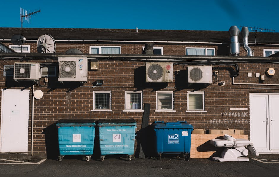 A row of three large waste bins positioned outside a brick building under a clear sky during daytime, with two blue recycling bins and one dark blue general waste bin placed on the pavement. The building's exterior features multiple air conditioning units and vent pipes mounted on the wall, along with small windows with white frames. A white door and a sign indicating a delivery area are visible on the right side of the building. Nearby, a stack of white plastic items and wooden pallets are arranged. The scene depicts the process of clearing bulky waste after a home relocation or moving, a service often provided by companies like Man with Van West Hendon, which specialises in removals, packing, and transporting furniture and belongings in West Hendon.