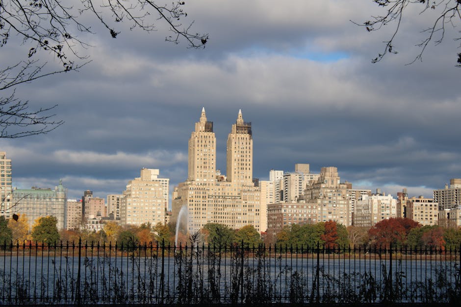 A wide view of the Manhattan skyline across a body of water, featuring two prominent high-rise buildings with pointed tops, surrounded by various other tall buildings of different architectural styles. In the foreground, a black metal fence runs horizontally along the water's edge, with some bushes and trees nearby, some with autumn foliage in orange and red hues. The sky overhead is partly cloudy with a mix of dark and light clouds, creating a moody atmosphere. The scene captures a typical cityscape core to professional home relocation and furniture transport services, reflecting the importance of logistics in packing and moving processes, as sometimes seen during house removals near key landmarks like Welsh Harp Reservoir. Man with Van West Hendon, specializing in removals, often coordinates loading and transportation work in such urban environments, ensuring smooth transit for belongings during home relocations.