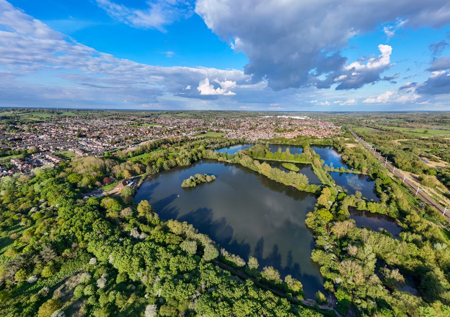 An aerial view of Welsh Harp Reservoir showing a large body of water surrounded by lush green parkland, walking paths, and scattered trees. In the foreground, a section of a property yard contains stacked cardboard boxes, wrapped furniture, and packing materials, possibly prepared for a home relocation at Man with Van West Hendon. A moving van is partially visible nearby, with furniture and boxes being loaded onto or off the vehicle using trolleys and straps. The scene is captured during daytime under partly cloudy skies, highlighting the space for furniture transport, packing, and logistics involved in house removals near Welsh Harp Reservoir.