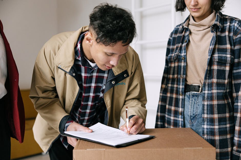 A man with curly dark hair, wearing a beige jacket over a checked shirt, is bent over a cardboard moving box, signing or filling out a document on a clipboard. Next to him stands a woman dressed in a beige turtleneck and a blue plaid shirt, with her hands resting by her sides. The setting appears to be indoors, likely within a residential property, with a white wall or shelving unit in the background. The scene is well-lit with natural or indoor lighting, highlighting the process of home relocation, packing, or moving preparations, which is consistent with services offered by Man with Van West Hendon, a professional removals company.