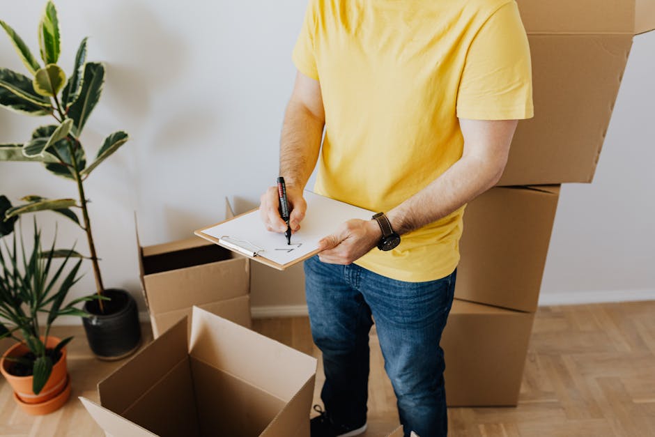 A man wearing a yellow t-shirt and blue jeans is standing indoors, surrounded by several large cardboard moving boxes, some open and empty, which are positioned on the light wooden floor near a white wall. He is holding a clipboard in one hand and writing with a black marker in the other, likely creating a packing or loading checklist as part of home relocation preparations. To his left, there is a tall potted plant with broad green leaves, adding indoor greenery to the space. The environment is well-lit, suggesting natural or bright indoor lighting, and the scene reflects the process of packing, planning, or inventory organization associated with professional removals, such as those offered by Man with Van West Hendon. This image visually supports content related to furniture transport and moving logistics within a residential setting, aligned with a headline about moving checklists for residents of West Hendon estate.