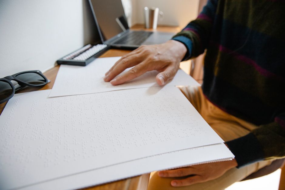 A person wearing a dark, long-sleeved shirt with colorful stripes, seated at a wooden desk, is handling large sheets of cardboard with braille dots, which are arranged for packing or preparing items f