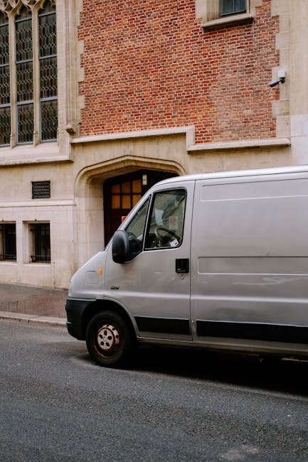 A light grey commercial van parked on a paved street in front of a historic brick building with large arched windows and a stone entrance. The van, which is positioned close to the building's entrance