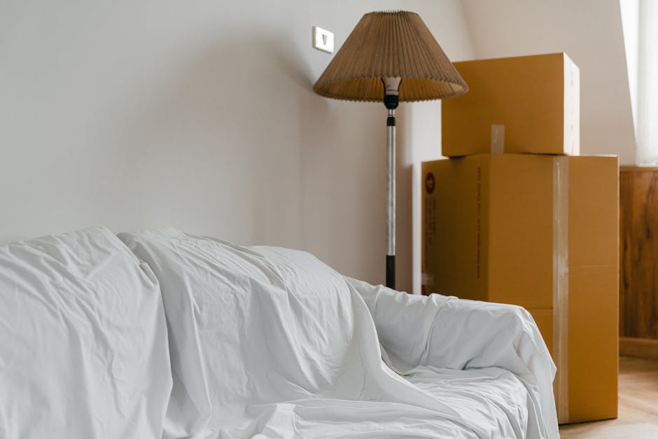 Image showing a living room with cardboard moving boxes stacked in the corner near a wooden cabinet. A beige floor lamp with a brown lampshade stands beside the boxes, casting light onto the plain light-colored wall. In the foreground, a white fabric-covered sofa is visible, positioned facing away from the boxes. The scene depicts an interior space prepared for packing and moving, with the boxes and furniture indicating a home relocation process, as might be arranged by Man with Van West Hendon during their house removals service.