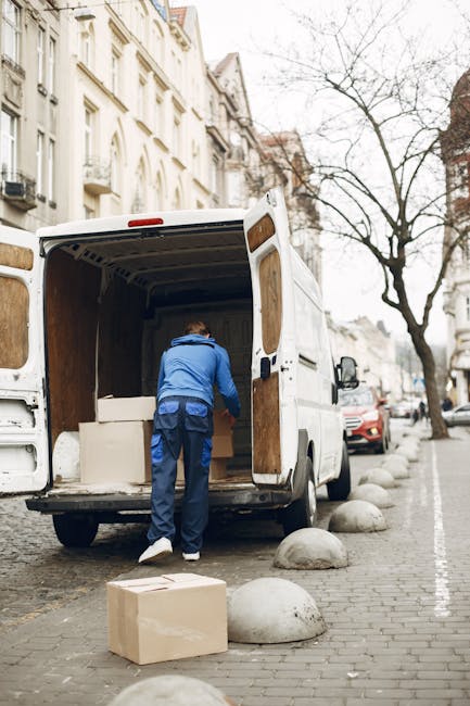 A man dressed in a blue jacket and dark trousers is loading a large cardboard box into the open cargo area of a white commercial van parked on a city street. The van, part of a house removal or furniture transport operation, has its rear doors fully open, revealing wooden panels lining the interior. Several other cardboard boxes are placed on the pavement nearby, ready for loading or unloading. The street is lined with multi-storey residential buildings with light-colored facades, and a leafless tree is visible in the background. The pavement features rounded stone bollards separating the walking area from parked vehicles, and a red car is parked close to the van. Overcast weather suggests natural daylight and typical urban surroundings for home relocation or packing and moving services, with the image capturing a moment within the loading process undertaken by Man with Van West Hendon.