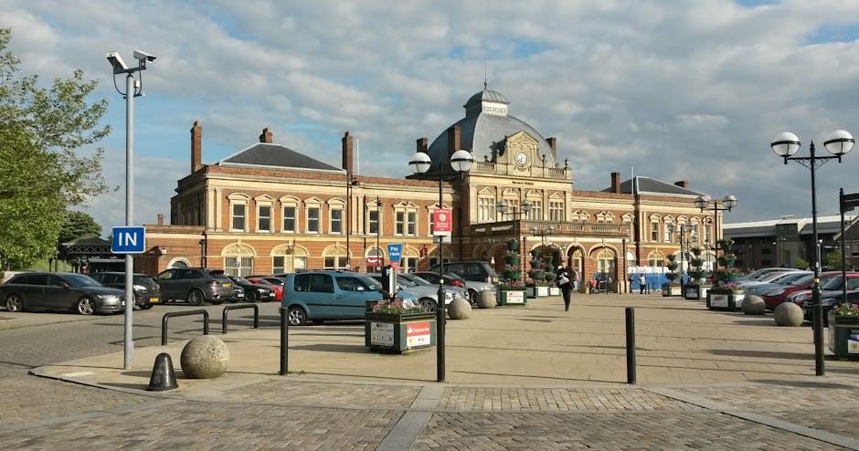 A large brick building with decorative architectural details and a clock on the central facade, situated behind a spacious paved area used for parking and loading. In the foreground, multiple parked cars are visible, with some near the building and others along the pavement. Several black metal bollards and spherical stone barriers demarcate the loading zone from pedestrian areas. Flower planters with blooming flowers add colour to the scene, positioned near street lamps with spherical lighting fixtures. A person is seen walking across the open space, possibly preparing for home relocation or furniture transport. The area is well-lit, with a partly cloudy sky overhead. The ambient environment suggests a public parking area suitable for house removals, with provisions for loading and vehicle manoeuvring, supporting efficient packing and moving logistics, as highlighted in the West Hendon Broadway parking tips page by Man with Van West Hendon.