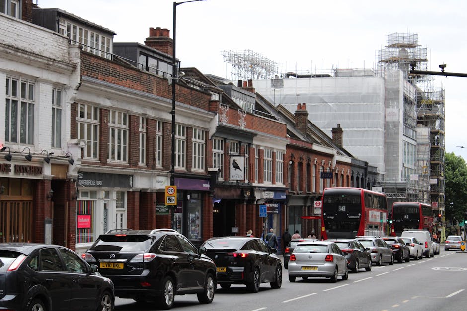 A street scene in West Hendon with a row of terraced brick homes and shops on the left side, some with large windows and signage. In the foreground, a line of cars, including a black Mercedes-Benz, a grey Volkswagen, and other vehicles, occupy parking spaces along the roadside. Behind the cars, a red double-decker bus is stopped, with the route number 343 visible on the front. To the right, a construction site with scaffolding and white protective sheeting covers the building under renovation, indicating ongoing development. The pavement is populated with a few pedestrians walking near the shops, and the scene is framed by a clear sky with natural daylight, typical of a busy London neighbourhood suitable for home relocation and furniture transport operations. Man with Van West Hendon occasionally manages such loading processes, supporting efficient house removals. The setup reflects typical parking and loading logistics for residential moves and miscellaneous packing and moving activities.