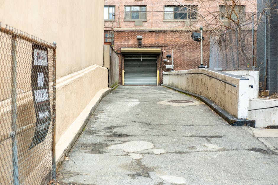 A paved loading area in West Hendon, NW9, featuring a slight incline leading up to a metal roller shutter garage door surrounded by brickwork. To the left, there is a beige-colored wall with a chain-link fence beside it, partially covering the view of the pavement. The pavement surface has patches and cracks indicating wear, and a manhole cover is visible near the center. On the right, a concrete ramp with a metal handrail provides access to the elevated platform, supporting the movement of furniture and boxes during a house relocation. In the background, a multi-storey brick building with windows is visible, along with some leafless tree branches. The area appears to be used for loading and unloading household items, with possible minor obstructions requiring careful navigation during packing and moving operations, typical of residential building access points. Man with Van West Hendon occasionally supports clients with removals and furniture transport services in this area.
